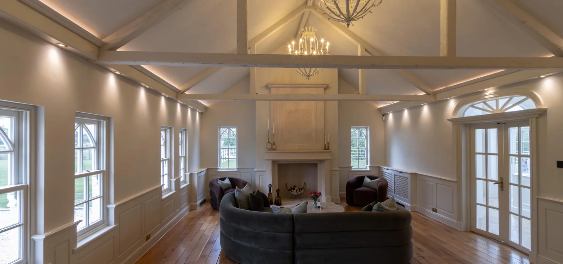 Large open-plan living space with a vaulted beamed ceiling, chandeliers, panelled walls and a central fireplace